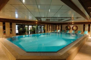 Rechteckiger Hotel-Innenpool mit türkisfarbenem Wasser, Liegestühlen und Pflanzen, umgeben von edlem Marmor und Holzverkleidung.