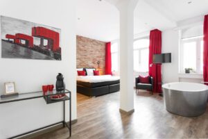 Doppelzimmer im 1st Floor Köln 2 mit roten Vorhängen, Backsteinwand, Sessel und freistehender Badewanne.